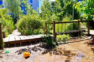 Hölzerne Brücke und schmale Pfade durch einen üppigen, naturnah gestalteten Spielbereich in einer Kindertagesstätte, umgeben von grünen heimischen Sträuchern und hellem Sonnenschein
