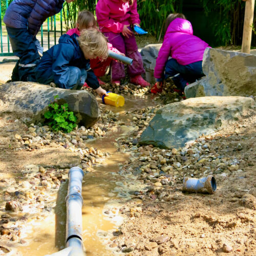 Gruppe von Kindern spielt zusammen an einem wassergefüllten Matschbach in einem naturnah gestalteten Außenbereich einer Kindertagesstätte.