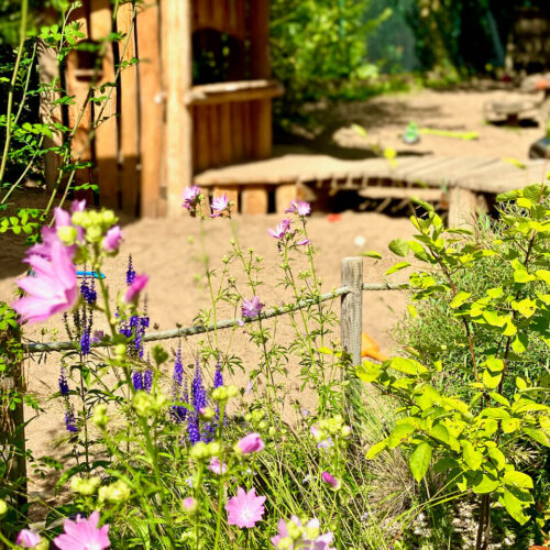 Verspielte Gartenansicht mit einem Spielhaus aus Robinienholz im Hintergrund, umgeben von blühenden Wildblumen und grünen Pflanzen