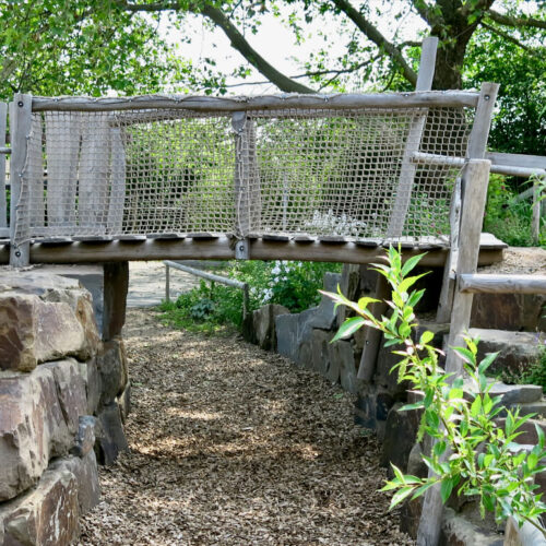 Rustikale Holzbrücke mit Netz überquert einen Weg in einem naturnah gestalteten Kindergarten, umgeben von blühenden Pflanzen und Sträuchern