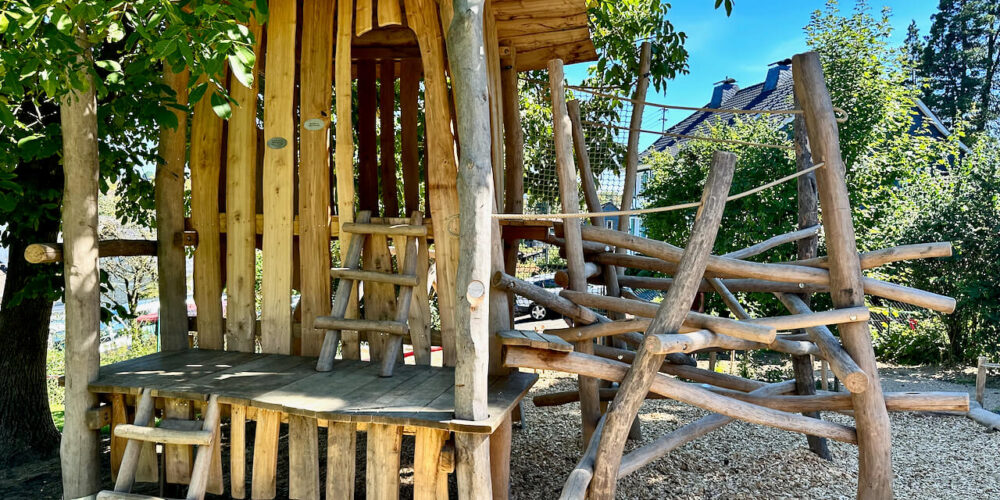 Holzspielhaus aus Robinie in einem naturnah gestalteten Außengelände einer Kindertagesstätte, umgeben von Grün, mit integriertem Klettermikado und Seilnetz unter sonnigem Himmel