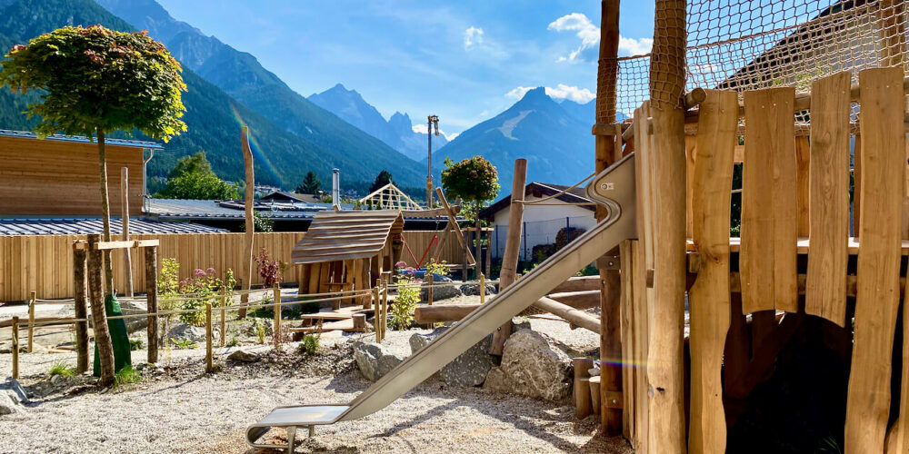 Kinderspielplatz mit Spielhütte aus Robinie, Schaukel und Rutsche vor einer beeindruckenden Bergkulisse unter klarem blauem Himmel