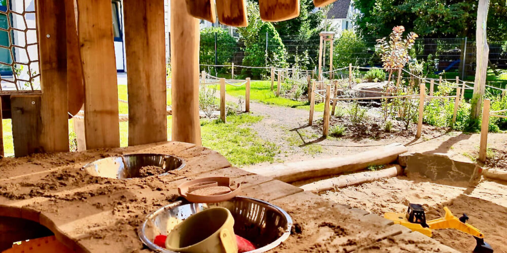 Blick aus Spielhütte mit Theke mit Sand und Spielzeugen im Vordergrund, mit einem naturnahen Garten und heimischen Pflanzen im Hintergrund